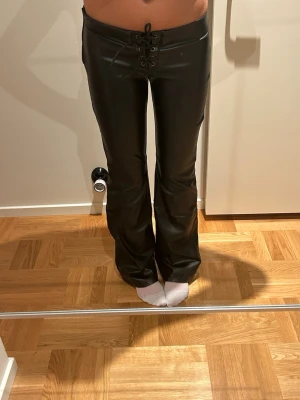 Svarta skinnbyxor från Zara - Säljer ett par svarta skinnbyxor från Zara med snörning framtill. Byxorna har en tight passform upptill och är utsvängda nedtill för en trendig look. Perfekta för dig som vill ha en edgy stil.