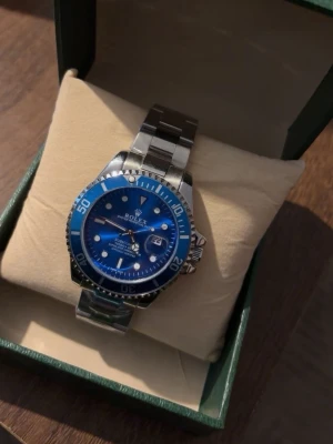 Klocka - Snygg klocka med blå urtavla och blå bezel. Klockan har ett robust armband i silverfärgat stål och tydliga indexmarkeringar. Klassisk design med datumfönster och vridbar bezel, perfekt för dig som gillar stilrena accessoarer.