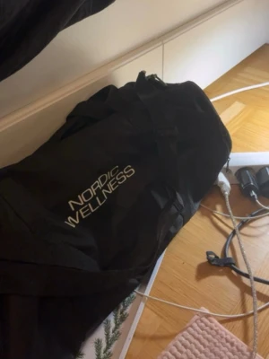 Svart träningsväska Nordic Wellness - Säljer en svart sportig träningsväska med Nordic Wellness-logga. Väskan har dubbla handtag och axelrem, perfekt för gymmet eller träning. Tillverkad i slitstarkt tygmaterial och har rymligt huvudfack.