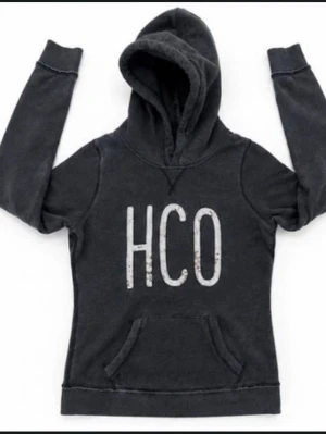 Mörkgrå hoodie med HCO-tryck - Snygg mörkgrå hoodie med stor huva och magficka framtill. På bröstet står det HCO i stora vita bokstäver. Hoodien har ribbade muddar vid ärmsluten och en avslappnad passform. Perfekt för en chill och casual stil.