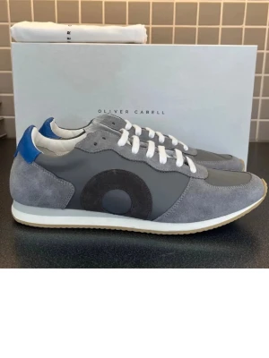Grå sneakers från Oliver Cabell - Snygga grå sneakers från Oliver Cabell med mockadetaljer, vit sula och vit snörning. Skorna har en svart cirkeldetalj på sidan och blå hälflik. Perfekta för en clean och modern stil.