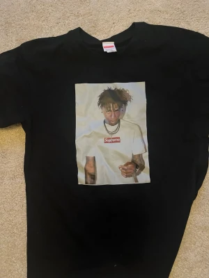 Svart t-shirt med Supreme-tryck - Säljer en svart t-shirt med ett stort tryck på framsidan som föreställer en person i vit Supreme-tröja. T-shirten har rund hals och korta ärmar. Perfekt för dig som gillar streetwear och vill sticka ut.