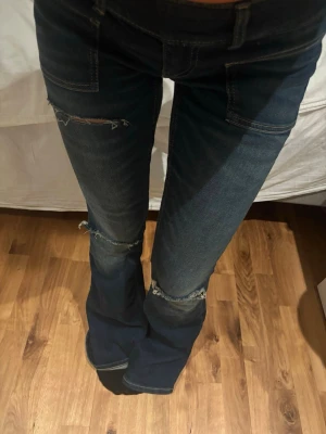 Mörkblå bootcut jeans med slitningar - Säljer ett par mörkblå bootcut jeans med slitna detaljer vid knäna och låret. Jeansen har låg midja, två knappar i linningen och klassiska fickor fram. Materialet är denim med stretch för skön passform. Perfekta för en trendig och avslappnad look.