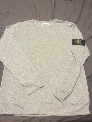 Grå sweatshirt från Stone Island - Snygg grå sweatshirt från Stone Island med klassisk rund hals och ribbade muddar. Tröjan har den ikoniska Stone Island-loggan på vänster ärm och är tillverkad i mjukt bomullsmaterial. Perfekt för en clean och avslappnad stil.