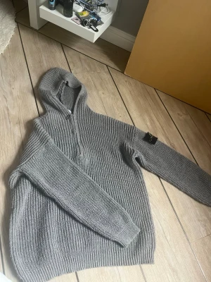 Grå stickad half zip hoodie från Stone Island - Säljer en grå stickad hoodie från Stone Island med dragkedja framtill och ribbad struktur. Tröjan har huva och långa ärmar samt den klassiska Stone Island-loggan på ärmen. Perfekt för dig som gillar streetwear och vill ha något både snyggt och bekvämt.