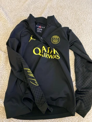 Svart PSG x Jordan träningströja - Snygg svart träningströja från PSG x Jordan med gul logga och detaljer. Halvlång dragkedja framtill, tryckt Paris Saint-Germain-märke på bröstet och Qatar Airways-tryck. Slim fit-modell med coola mönster på ärmarna. Perfekt för fotbollsträning eller streetwear.