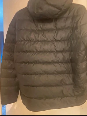 Svart dunjacka från Nike Therma-Fit - Snygg svart dunjacka från Nike med Therma-Fit-teknologi. Jackan har huva och quiltad design med breda kanaler. Perfekt för kalla dagar när du vill hålla värmen och ändå se stilren ut.