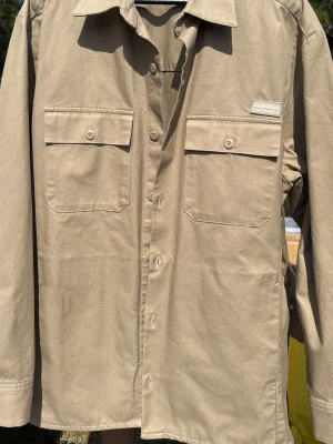 Beige overshirt från J.Lindeberg - Stilren beige overshirt från J.Lindeberg i bomull. Två stora bröstfickor med lock och knappar framtill. Klassisk krage och raka ärmar. Perfekt lager-på-lager-plagg för en avslappnad look.
