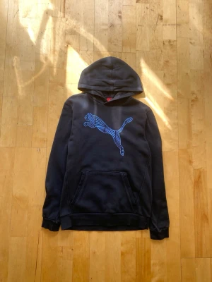Svart hoodie med blå Puma-logga - Säljer en svart hoodie från Puma med stor blå logga på bröstet. Hoodien har huva och en klassisk magficka. Tillverkad i mjukt bomullsmaterial och har ribbade muddar vid ärmslut och nederkant. Perfekt för en avslappnad och sportig stil.