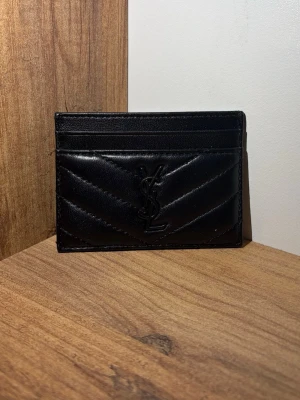 Svart quiltad YSL kortfodral - Snyggt svart kortfodral i quiltat skinn med klassisk YSL-logga framtill. Smidig och stilren design med flera kortfack, perfekt för dig som vill ha ett lyxigt och kompakt alternativ till plånbok.
