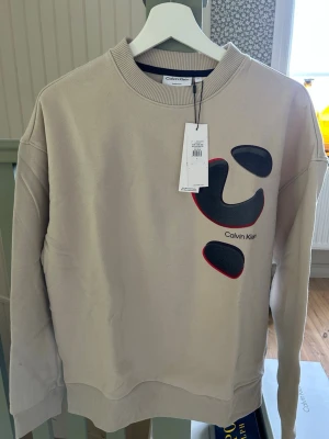 Beige sweatshirt från Calvin Klein - Säljer en beige sweatshirt från Calvin Klein med svart och rött abstrakt tryck på bröstet. Oanvänd och nypris 1399kr