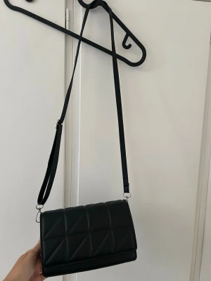 Svart quiltad axelväska - Snygg svart axelväska i quiltad design med justerbar axelrem. Väskan har magnetlås och ett rymligt huvudfack. Tillverkad i konstläder med stilrena silverdetaljer. Perfekt för dig som gillar en clean och modern look.