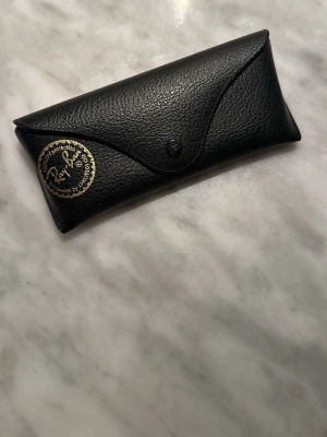 Svart Ray-Ban solglasögonfodral - Snyggt svart fodral från Ray-Ban i strukturerat skinn, perfekt för att skydda dina solglasögon. Klassisk design med tryckknapp och Ray-Ban-logga i guld på framsidan. Passar till de flesta modeller av solglasögon. Ink orginal putsduk. 