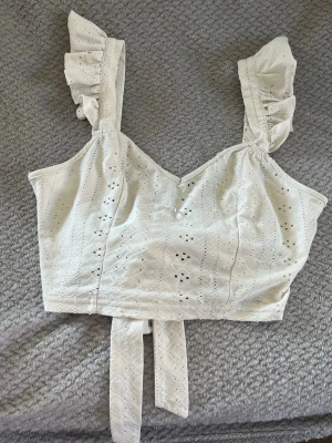 Vit broderad linnetopp med volang - Söt vit linnetopp med broderade detaljer och volangprydda axelband. Toppen har v-ringning och knytband i ryggen för en extra fin touch. Perfekt för varma dagar och ger en romantisk vibe till din outfit.