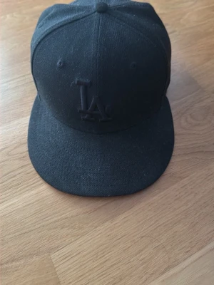 Svart LA-keps från New Era 59FIFTY - Säljer en svart keps från New Era 59FIFTY med LA-logga framtill. Klassisk flat brim och broderad logga i svart för en clean look. Tillverkad i bomull och polyester, perfekt för dig som gillar streetwear och sportig stil.