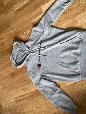 Grå hoodie från Champion - Snygg grå hoodie från Champion med stor logga i blått och rött på bröstet. Klassisk känguruficka framtill och justerbar huva med dragsko. Tillverkad i mjuk bomullsblandning, perfekt för en avslappnad streetwear-look.