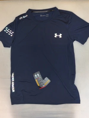 Blå tränings-tshirt från Under Armour - Snygg mörkblå tränings-tshirt från Under Armour med logga på bröstet och tryck på ärmen och sidan. Tillverkad i ett lätt och snabbtorkande funktionsmaterial som passar perfekt till gymmet eller löprundan. Klassisk passform och rund hals.