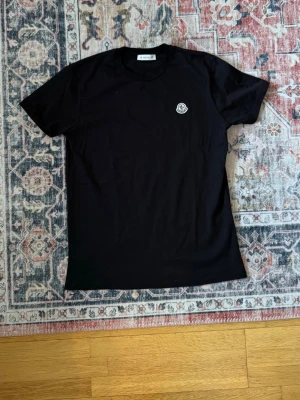 Svart Moncler T-shirt - Säljer en svart t-shirt från Moncler med klassisk passform och Moncler-logga på bröstet. Tillverkad i mjuk bomull och har rund hals samt korta ärmar. Perfekt för en clean och stilren look. Går att skanna NFC och sitter som S