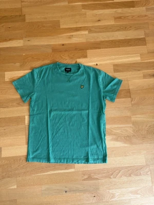 Grön t-shirt från Lyle&scott - Snygg grön t-shirt med rund hals och korta ärmar. På bröstet sitter en liten broderad logga i gult och svart som ger en clean look. Perfekt till jeans eller shorts för en avslappnad stil.