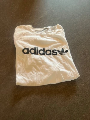 Vit Adidas t-shirt med logga - Säljer en klassisk vit t-shirt från Adidas med stor svart logga och text på bröstet. T-shirten har rund hals och korta ärmar, perfekt till en avslappnad stil. Materialet känns mjukt och skönt mot huden.