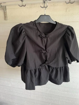 Svart puffärmad blus från SHEIN - Trendig svart blus från SHEIN med puffiga trekvartsärmar och dekorativa knytningar framtill. Blusen har rund halsringning och volang nedtill, vilket ger en söt och modern look. Perfekt för dig som gillar statement-plagg och vill sticka ut.