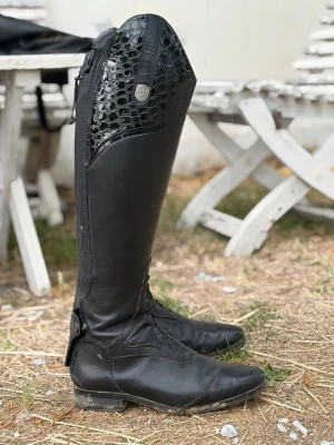 Svarta ridstövlar (dressyr) - Säljer ett par höga svarta ridstövlar i läder med snörning framtill och dragkedja bak. Stövlarna har en snygg krokopräglad detalj upptill och en liten metallbadge på sidan. Perfekta för dig som vill ha en stilren och klassisk look i stallet. De kostar 4000 nya men säljer för 2700. De är använda max 3 gånger men används inte i nuläget. De är från mountain horses  
