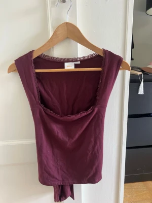 Vinröd ärmlös topp med knyt - Säljer en vinröd ärmlös topp med snygg knytning i ryggen och spetsdetaljer vid halsringningen. Toppen har en croppad passform och är tillverkad i mjuk bomull, perfekt för att lyfta vilken outfit som helst.