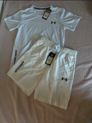 Vitt träningsset från Under Armour - Snyggt vitt träningsset från Under Armour med t-shirt och shorts. T-shirten har logga på bröstet och tryck på ärmen, medan shortsen har resår i midjan och logga på benet. Perfekt för gymmet eller löpning. Materialet känns lätt och andas bra.