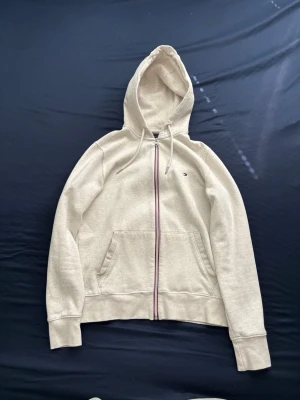 Tommy Hillfiger hoodie med dragkedja - Säljer en beige TH hoodie med dragkedja och huva. Hoodien har två fickor framtill och en liten broderad logga på bröstet. Mjuk bomullsblandning och ribbade muddar vid ärmar och nederkant. Perfekt för en avslappnad stil.