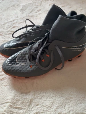 Grå Nike Hypervenom fotbollsskor - Snygga grå Nike Hypervenom fotbollsskor med högt skaft och snörning. Skorna har orange detaljer på sulan och vita loggor. Tillverkade i syntetmaterial för bra grepp och kontroll på planen. Perfekta för dig som vill sticka ut på matchen.