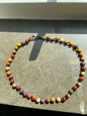 Färgglatt pärlhalsband i sten - Unikt halsband med runda pärlor i olika färger som gul, röd, lila, beige och brun. Pärlorna är troligen av sten och har naturliga mönster. Halsbandet har ett metallspänne och känns både retro och bohemiskt. Perfekt för dig som gillar färg och vill sticka ut.