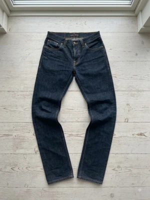 Nudie Jeans Grim Tim  - Snygga mörkblå jeans i rak modell med klassiska kontrastsömmar och fem fickor. Baksidan har två fickor med vågformad detaljsöm och en läderpatch i midjan. Jeansen är tillverkade i kraftig denim och har en tidlös look.