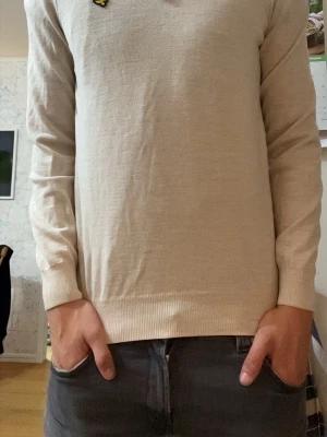  Beige lyle & scott  stickad tröja med halv dragkedja - Snygg beige stickad tröja från Lyle & Scott med halv dragkedja och gul logga på bröstet. Tröjan har lång ärm, ribbade muddar och en mjuk, tunn stickning som passar perfekt till lager på lager. Klassisk och stilren design.