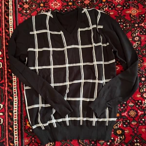 Svart stickad tröja med rutmönster - Snygg svart stickad tröja med vitt rutmönster på framsidan och V-ringning. Tröjan har långa ärmar och ribbade muddar vid ärmslut och nederkant. Perfekt för dig som gillar klassiska men trendiga plagg.