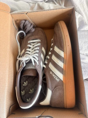 Adidas Spezial bruna sneakers - Bruna Adidas Spezial sneakers i mocka med vita detaljer och klassiska tre ränder på sidan. Skorna har vit sula och vita skosnören samt gummisula i brun ton. Låg modell med retrovibbar och Adidas-logga på plösen och innersulan. Helt nya med kartong!