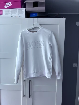 Vit sweatshirt från Hugo Boss - Snygg vit sweatshirt från Hugo Boss med stort BOSS-tryck på bröstet. Tröjan har rund halsringning, långa ärmar och är gjord i mjukt bomullsmaterial. Perfekt för en clean och stilren look.