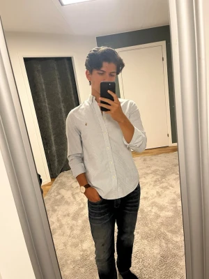 Randig skjorta från Ralph Lauren - Säljer en klassisk vit skjorta med tunna blå ränder från Ralph Lauren. Skjortan har button-down krage, långa ärmar och den ikoniska broderade loggan på bröstet. Perfekt till jeans eller chinos för en clean och stilren look. Kolla sista bilden det fins två små fläckar men de syns inte om du viker upp skjortan eller har något över som en tjocktröja. Fläckarna går säkert bort med rätt tvättmedel😊