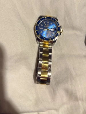 blå och guld klocka - Snygg klocka med blå urtavla och gulddetaljer. Armbandet är i silver och guldtonat metall, och boetten har en blå ring med guldfärgade markeringar. Klockan har klassisk länkdesign och tydliga indexmarkeringar.
