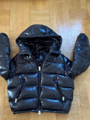 Zara jacka  - Snygg svart pufferjacka från Zara med glansig finish och stor huva. Jackan har två dragkedjeförsedda fickor framtill och breda, vadderade paneler som ger en riktigt puffig look. Perfekt för kalla vinterdagar och riktigt trendig stil.