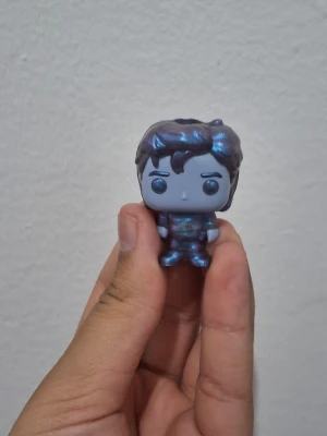 Blå-lila Funko Pop figur - Säljer en liten Funko Pop-inspirerad figur i blå-lila metallic färg. Figuren har stort huvud, markerade ögonbryn och detaljerat hår. Tillverkad i plast och har en glansig yta som skiftar i färg beroende på ljuset. Perfekt som dekoration eller samlarpryl.