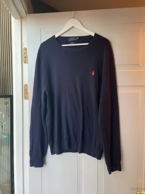 Mörkblå tröja från Polo Ralph Lauren - Klassisk mörkblå tröja från Polo Ralph Lauren med rund halsringning och den ikoniska röda logotypen broderad på bröstet. Tröjan har långa ärmar och ribbade muddar vid ärmslut och nederkant. Perfekt för en stilren och enkel look.