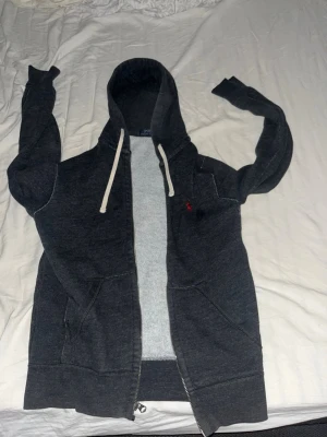 Mörkgrå hoodie från Polo Ralph Lauren - Mörkgrå hoodie med dragkedja från Polo Ralph Lauren. Klassisk design med vit dragsko i huvan och röd logga broderad på bröstet. Mjuk insida och stora fickor framtill. Perfekt för en chill och avslappnad stil.
