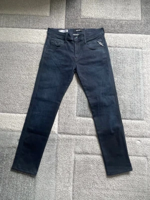 Mörkblå anbass slim fit jeans från Replay - Snygga mörkblå slim fit anbass jeans från Replay med klassisk femficksdesign. Jeansen har dragkedja och knapp framtill samt detaljerad söm på fickorna. Perfekta för en trendig och avslappnad stil.