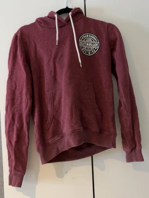 Vinröd hoodie Klitmøller med tryck - Säljer en vinröd hoodie från Klitmøller med stort vitt tryck på ryggen och mindre tryck på bröstet. Hoodien har huva med vita snören och en stor magficka. Tillverkad i mjukt bomullsmaterial, perfekt för chill dagar eller när du vill ha en avslappnad stil.