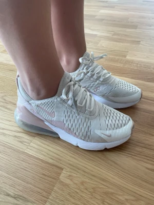 Nike Air Max 270 vita sneakers - Snygga Nike Air Max 270 sneakers i vitt med ljusrosa detaljer och genomskinlig luftbubbla i hälen. Ovandelen är i mesh för bra ventilation och skon har klassisk snörning. Perfekta för dig som gillar sportig och trendig stil.