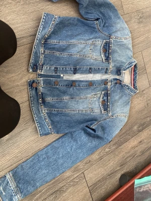 Klassisk blå jeansjacka - Snygg blå jeansjacka i klassisk modell med bruna knappar och två bröstfickor med lock. Jackan har raka sömmar och en lätt tvättad look som ger en avslappnad vibe. Perfekt att slänga över en hoodie eller t-shirt för en cool streetstyle.