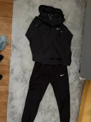 Svart Nike träningsset med huva nike runner - Säljer ett svart träningsset från Nike med långärmad jacka med huva och byxor. Jackan har dragkedja framtill och diskreta vita Nike-loggor på både jacka och byxor. Perfekt för träning eller löpning.