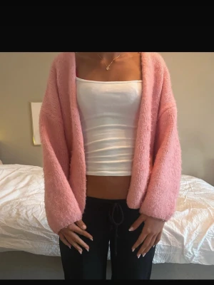 Ljusrosa fluffig kofta - Mysig ljusrosa kofta med öppen front och långa, vida ärmar. Koftan är fluffig och har en avslappnad passform, perfekt att slänga över en topp. Passar dig som gillar en soft och chill stil.