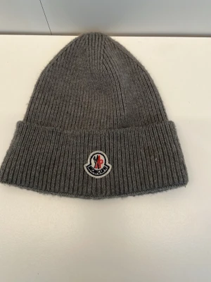 Grå ribbstickad mössa f - Säljer en grå ribbstickad mössa  med uppvikt kant och klassisk Moncler-logga framtill. Mössan är tillverkad i 70% ull och 30% kaninhår, vilket gör den extra mjuk och varm. Tillverkad i Italien och perfekt för kyliga dagar.