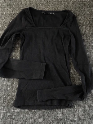 Svart ribbad långärmad topp - En svart, ribbad långärmad topp med fyrkantig ringning. Toppen har en slim passform och är tillverkad i ett mjukt, stretchigt material som passar perfekt till många olika stilar.